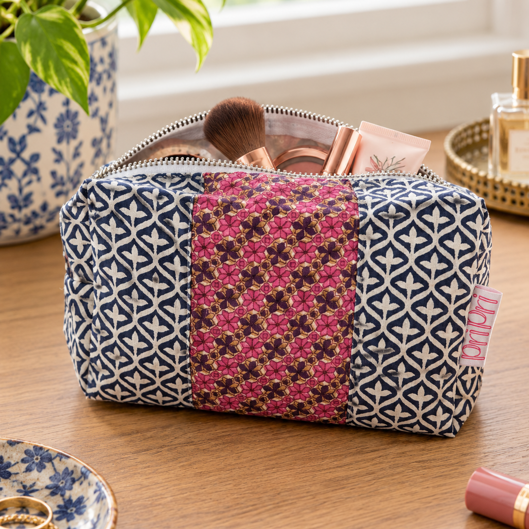 A fabric makeup bag with a navy blue geometric pattern and a central pink floral panel, shown on a wooden table with cosmetics and a plant nearby.