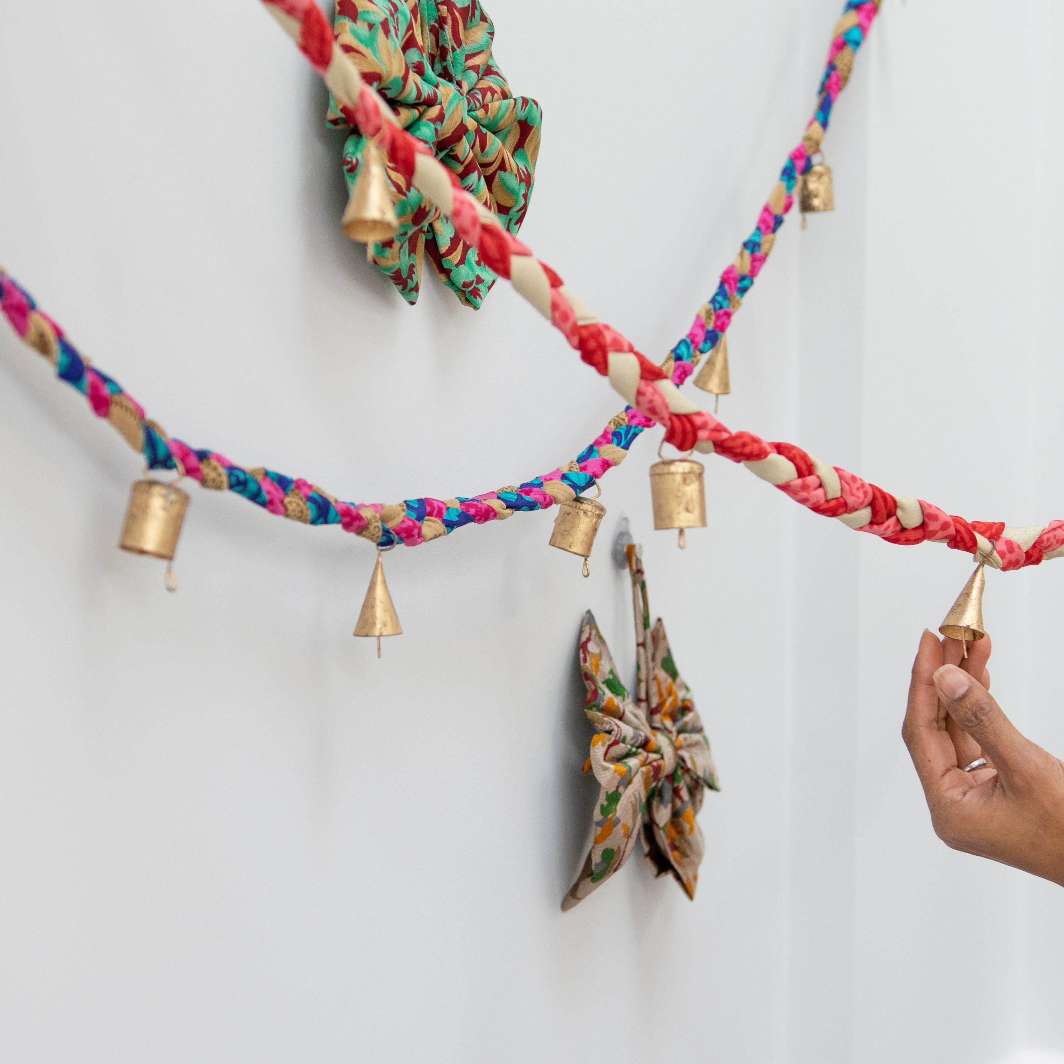 Colorful braided yarn garland with decorative bows and small gold bells on a white background.