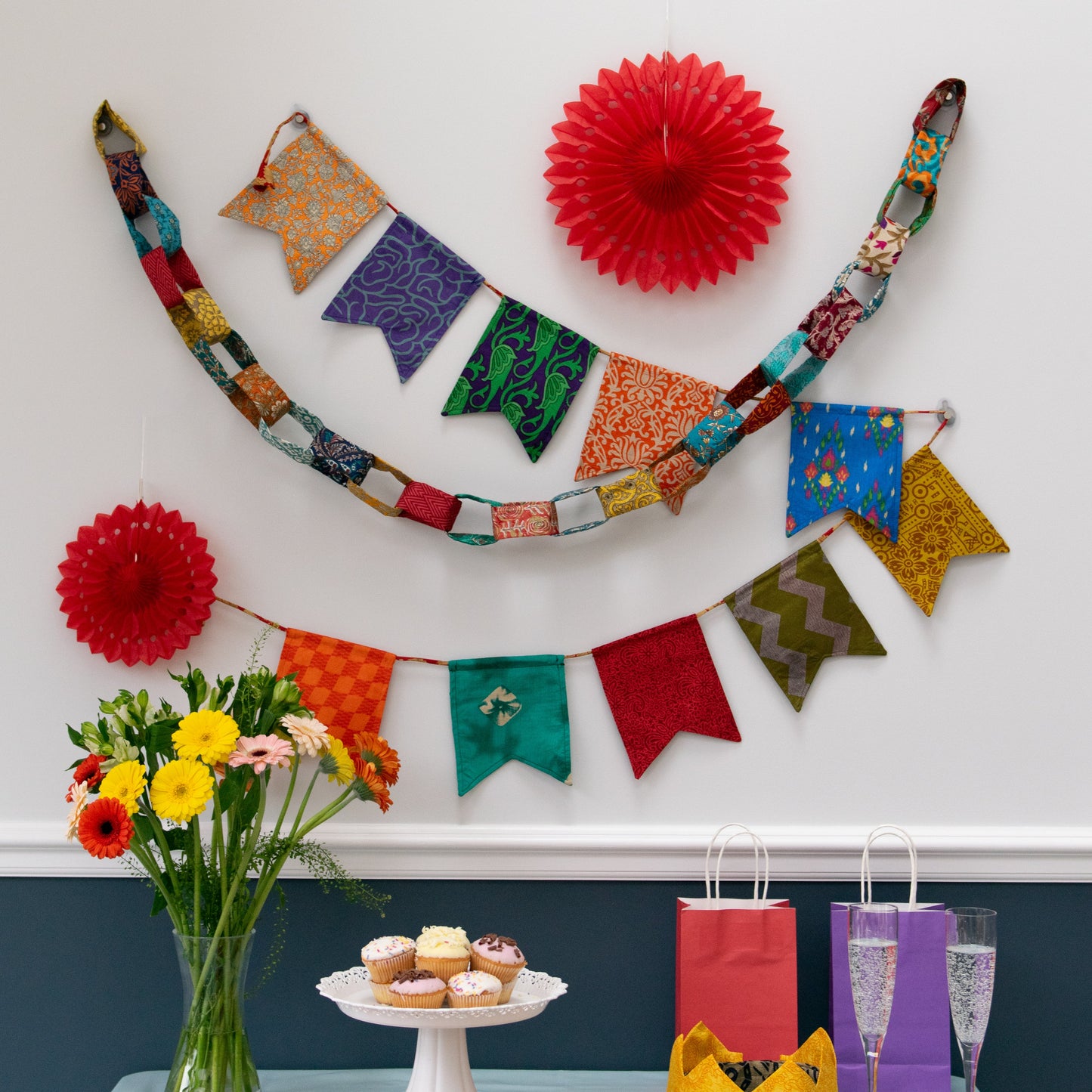 Decorative setup with colorful flags, flowers, and a cake stand on a table against a white wall.