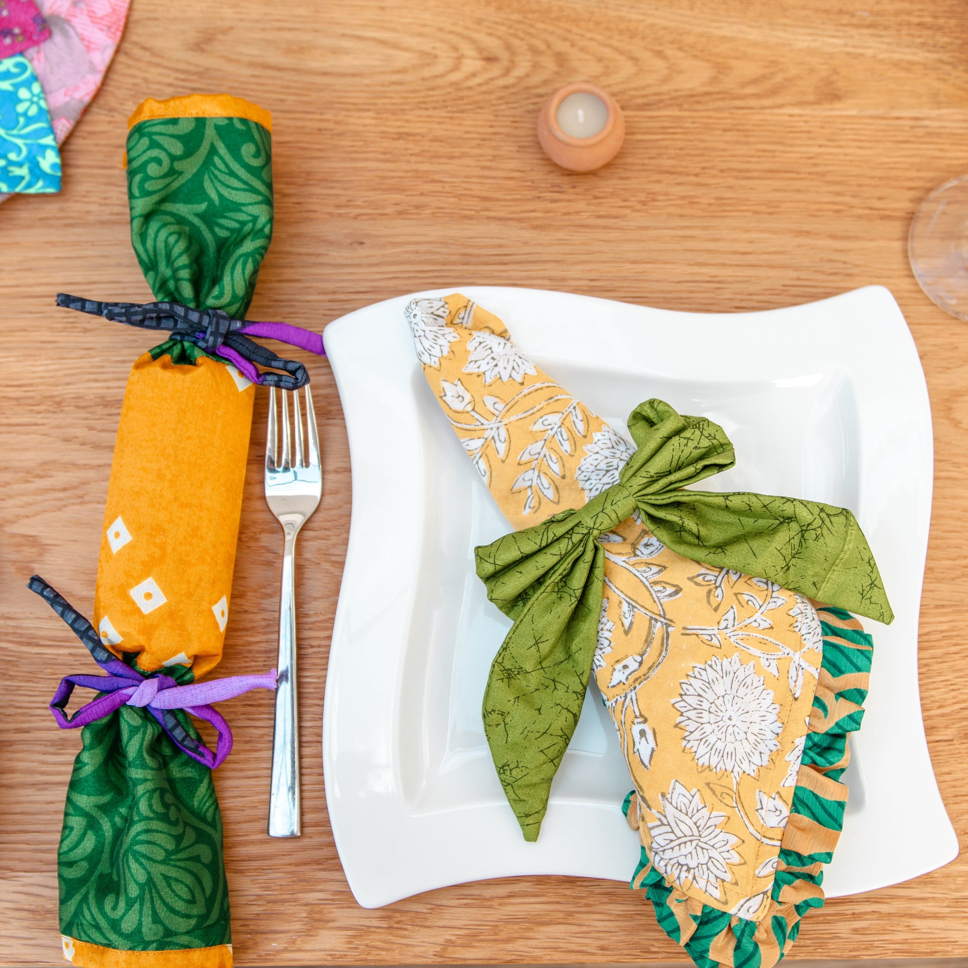 Decorative napkins and a fabric cracker tied with ribbons on a wooden table with cutlery and candles.