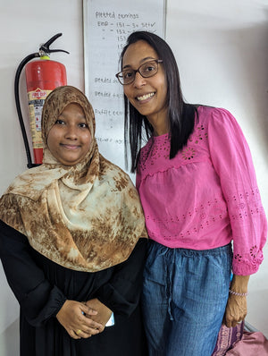 Two women standing together in an indoor setting. The woman on the left wears a black abaya and a brown patterned hijab, while the woman on the right wears a bright pink eyelet blouse and blue jeans.