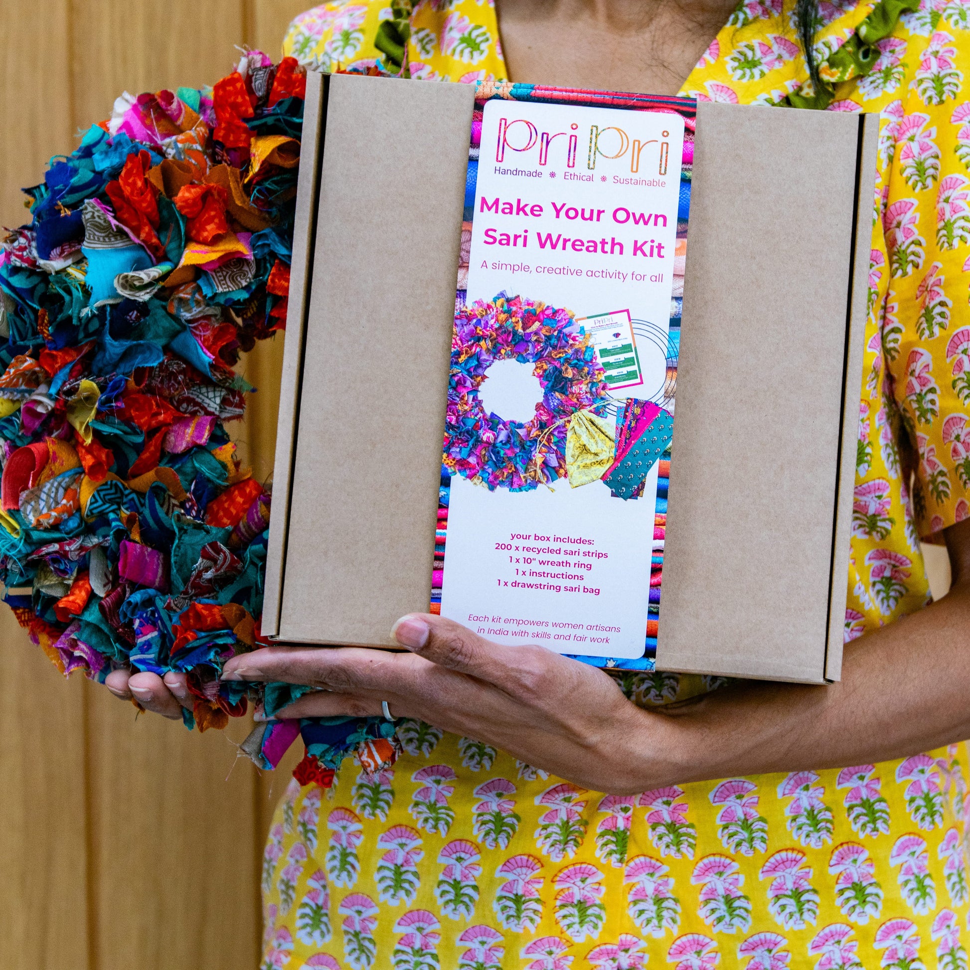 Person holding a colorful wreath and a 'Make Your Own Wreath Kit' by PriPri against a wooden background.
