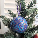 A round blue and purple Christmas ornament with gold floral patterns hanging from a green artificial Christmas tree branch.