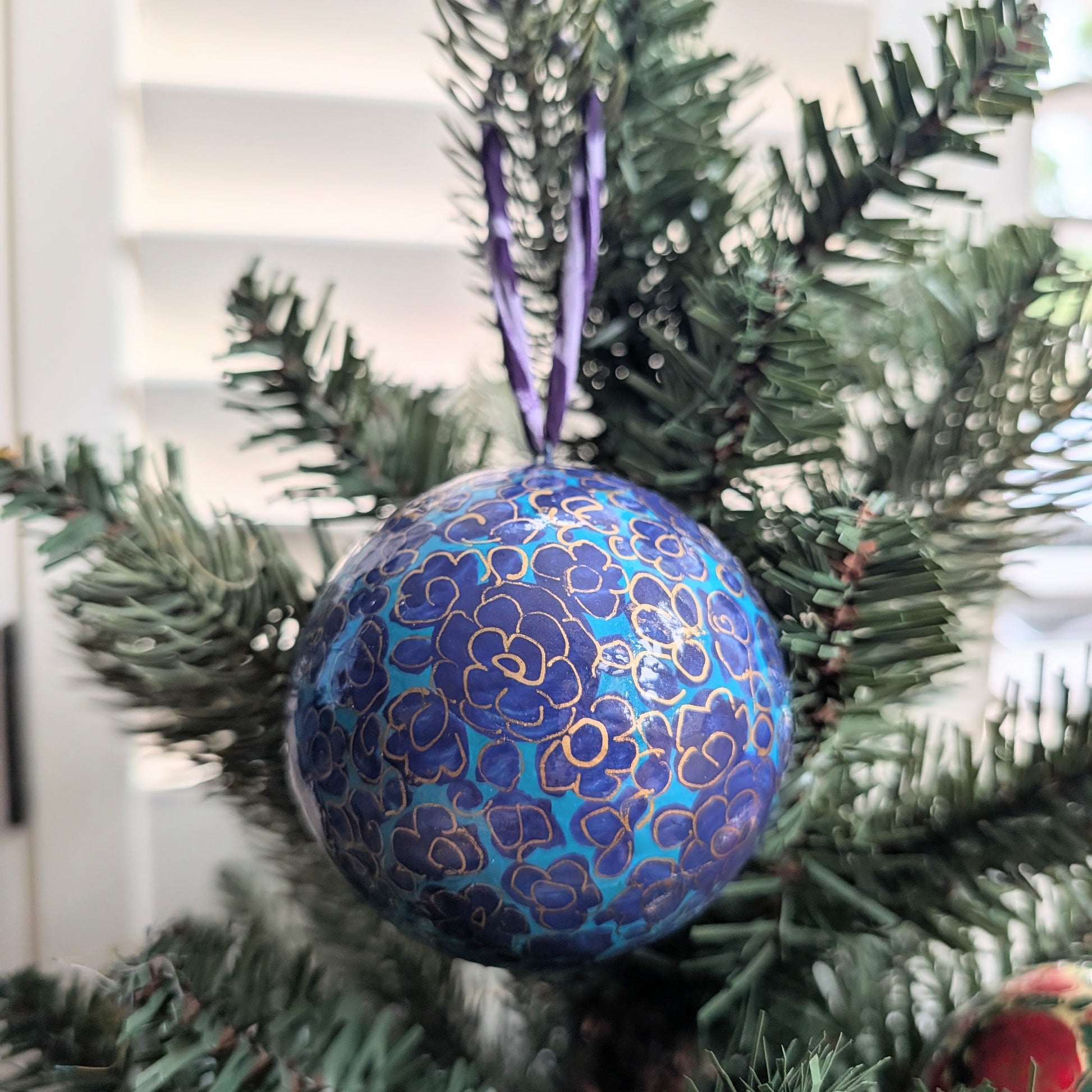 A round blue and purple Christmas ornament with gold floral patterns hanging from a green artificial Christmas tree branch.