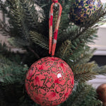 A red Christmas ornament with gold floral patterns and a red-and-white striped hanging ribbon, displayed on a green pine tree next to another patterned ornament.