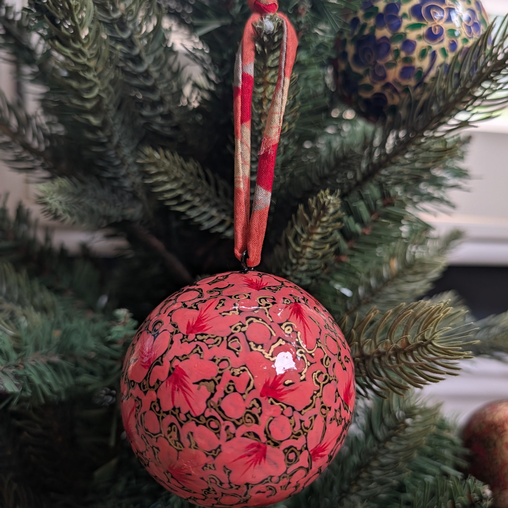A red Christmas ornament with gold floral patterns and a red-and-white striped hanging ribbon, displayed on a green pine tree next to another patterned ornament.