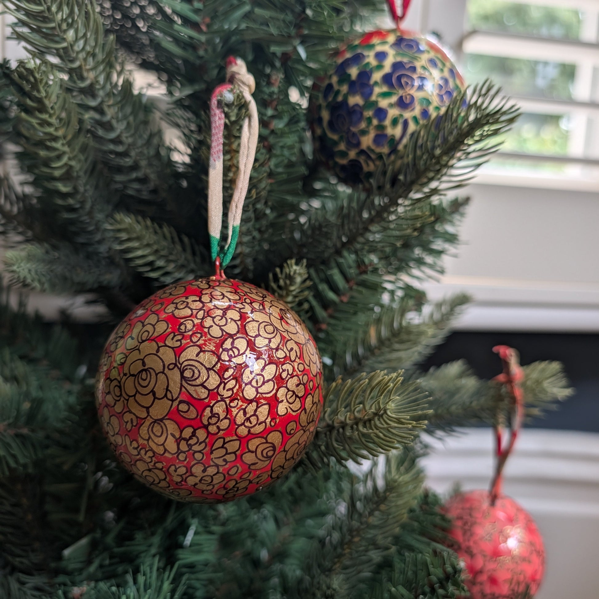 A close-up of a decorated Christmas tree with three hand-painted ornaments. The most prominent ornament in the foreground is a round red ball with gold floral patterns and a white hanging string.