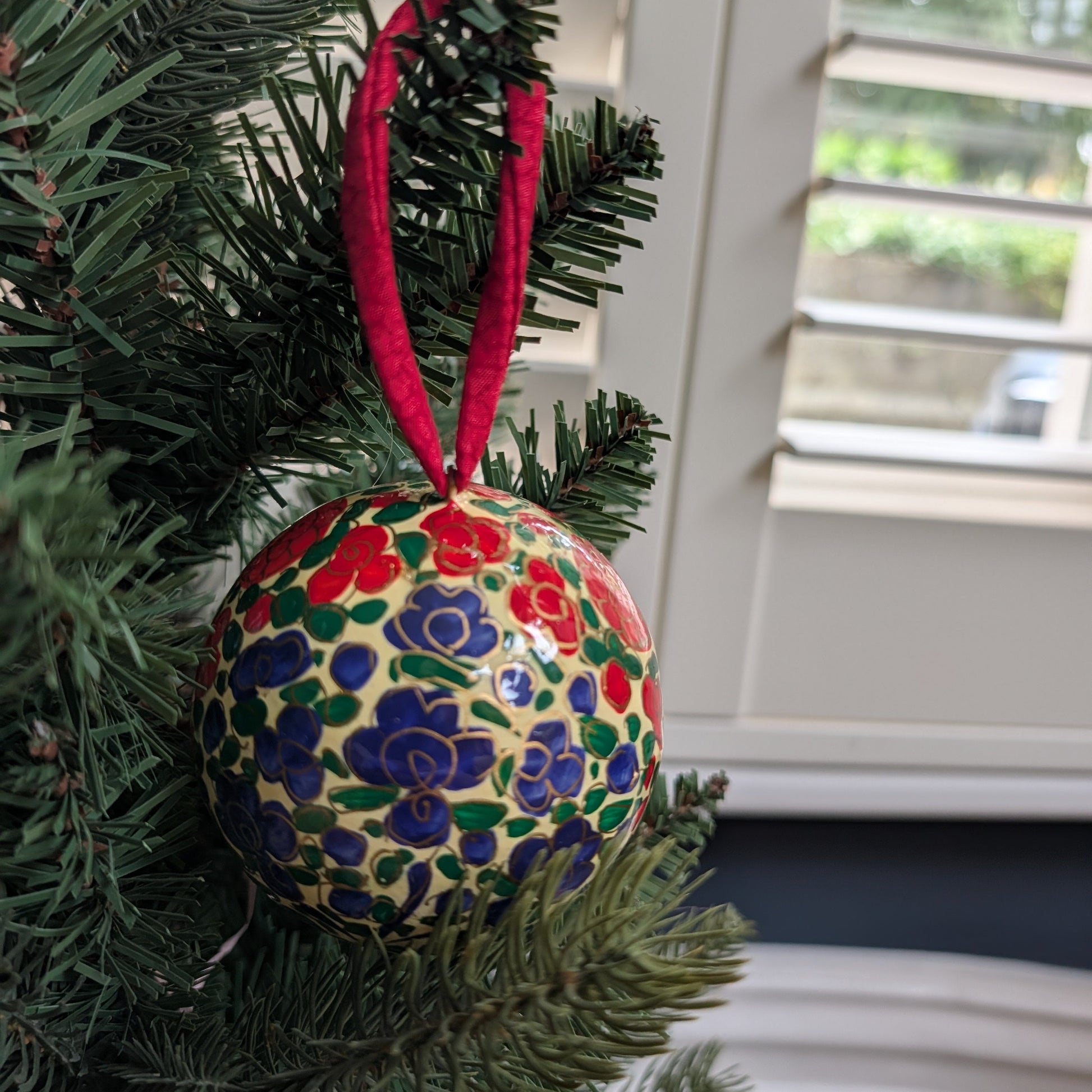 A hand-painted glass Christmas ornament with a red hanging ribbon, decorated with red, blue, green, and yellow floral patterns on a cream background, hanging on a green Christmas tree branch.