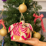 A hand holds a small gift box wrapped in a bright pink, red, and beige patterned fabric bow, set against a decorated Christmas tree with gold ornaments.