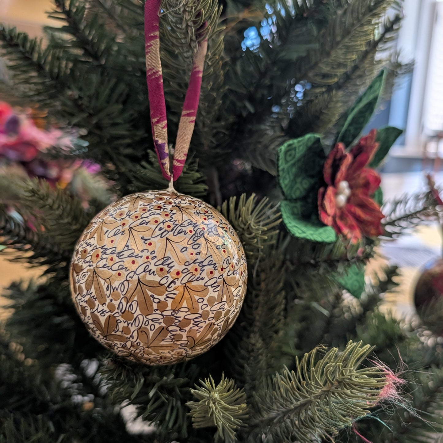 A close-up of a hand-painted gold Christmas ornament hanging on a green Christmas tree, with a patterned burgundy and beige ribbon.